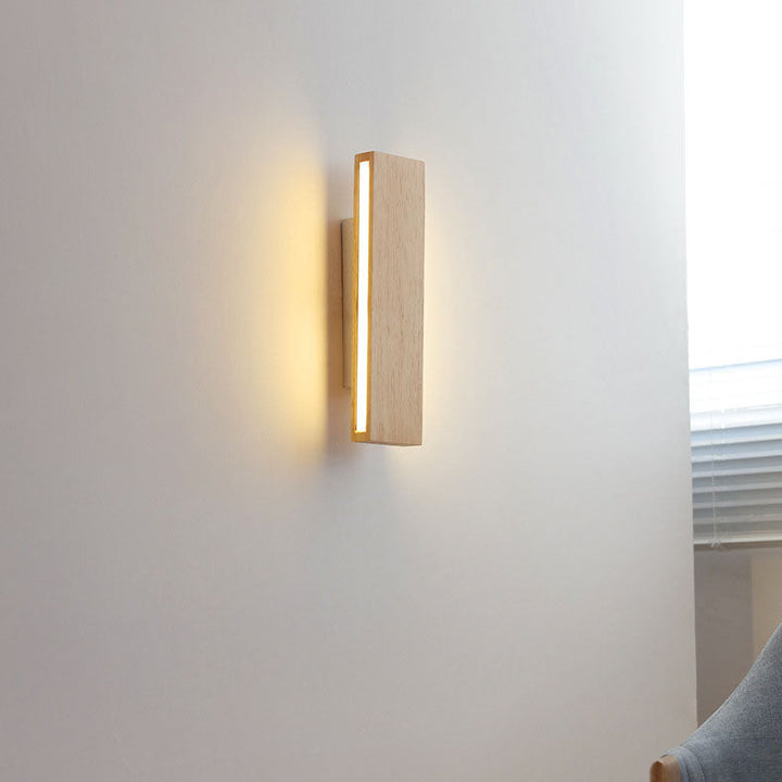 Verticale houten wandlamp aan een witte muur, brandend met warm licht, naast een raam en een grijze stoel. 15
