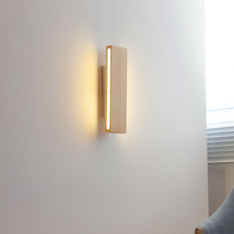 Verticale houten wandlamp aan een witte muur, brandend met warm licht, naast een raam en een grijze stoel. 15