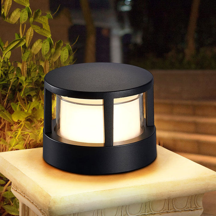 Ronde zwarte lantaarnlamp met plat dak en warm licht, 's avonds op een stenen sokkel naast groene planten. 1
