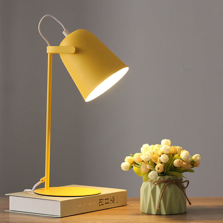 Gele bureaulamp met brandende kap op een boek, naast een groen vaasje met witte en gele roosjes. 1