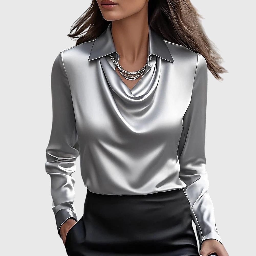 Model draagt de glanzende satijnen blouse in zilvergrijs met gedrapeerde hals en kraag, gecombineerd met een zwarte rok en strassketting. 2