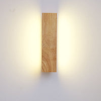 Frontaal zicht op de houten lamp in verticale stand, met warm licht dat links en rechts langs de muur schijnt. 3