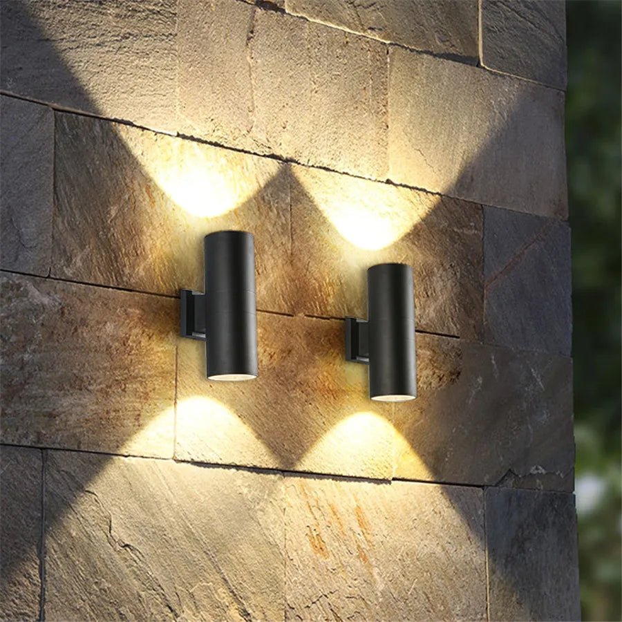 Twee zwarte cilinderlampen op een muur van natuursteen werpen warm licht omhoog en omlaag in een waaiervorm. 2