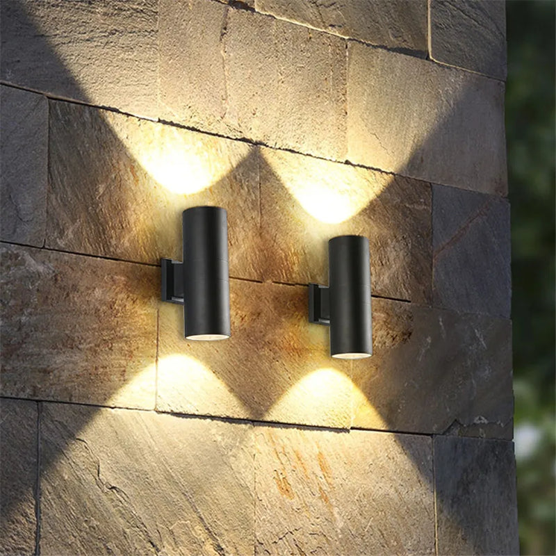 Twee zwarte cilinderlampen op een muur van natuursteen werpen warm licht omhoog en omlaag in een waaiervorm. 2
