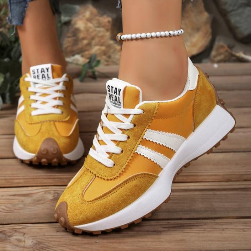 Gele suède sneakers met witte strepen aan de voeten van een model op een houten vloer 6