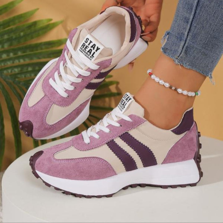Roze en beige sneakers met paarse strepen, één omhoog gehouden terwijl het model op een witte sokkel staat 11