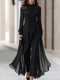 Model in zwarte chiffon tuniek met opstaande kraag en gedrapeerde sjaal over een zwarte broek, vanaf het hoofd tot de knieën gezien. 1