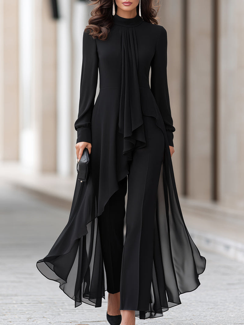 Model in zwarte chiffon tuniek met opstaande kraag en gedrapeerde sjaal over een zwarte broek, vanaf het hoofd tot de knieën gezien. 1