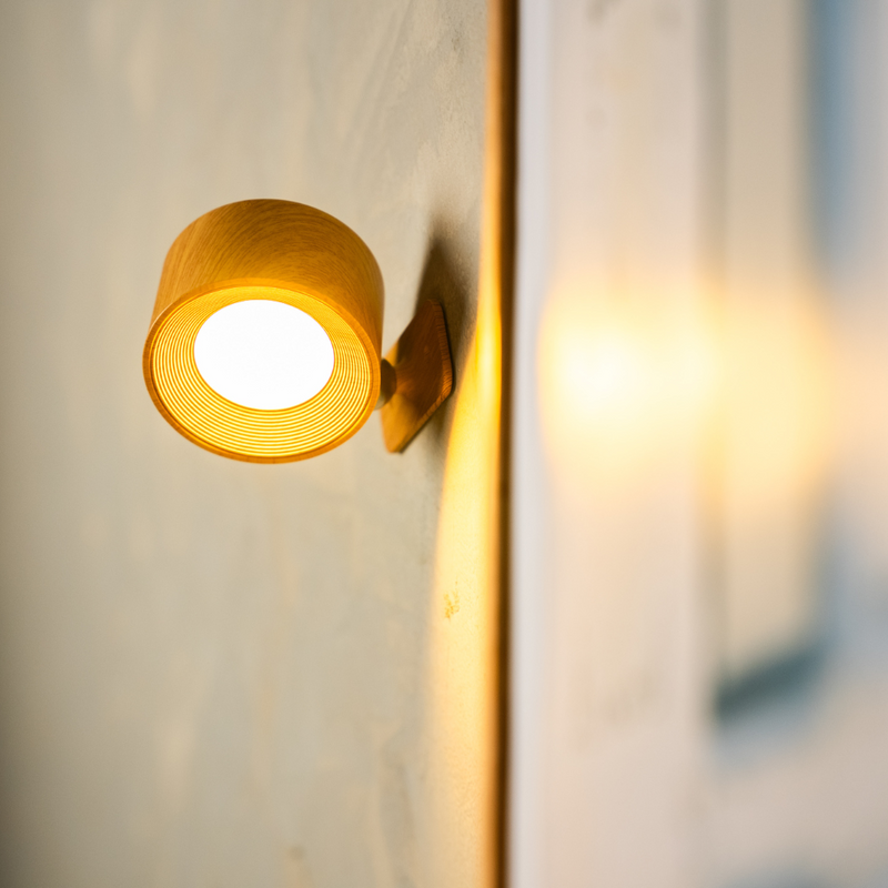 Close-up van de houtlook wandlamp met licht aan, met fijne ribbels rond de lichtbron en een houten montageplaat. 15