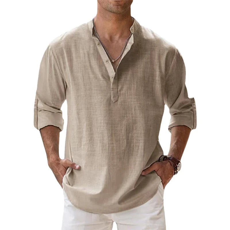 Man draagt het losvallende henleyshirt in beige met opgerolde mouwen en handen in de zakken van een witte broek. 1