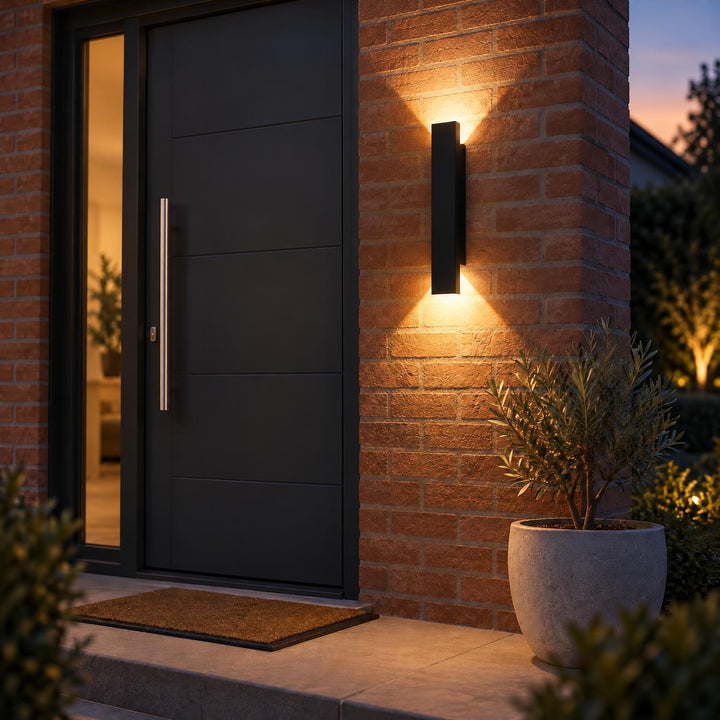Zwarte wandlamp op bakstenen muur bij de voordeur in de schemering, naast een plant in grijze pot 2