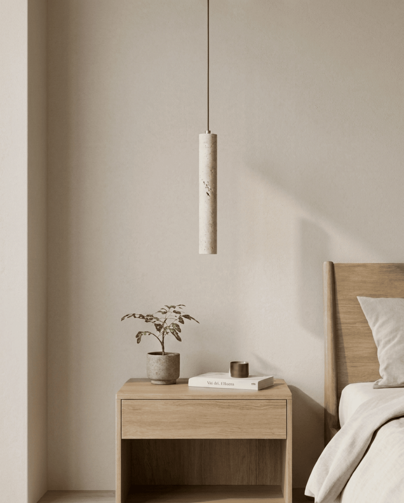 Uitgeschakelde cilindervormige travertijnen hanglamp boven een houten nachtkastje met plantje, boek en kaarsje in een 1