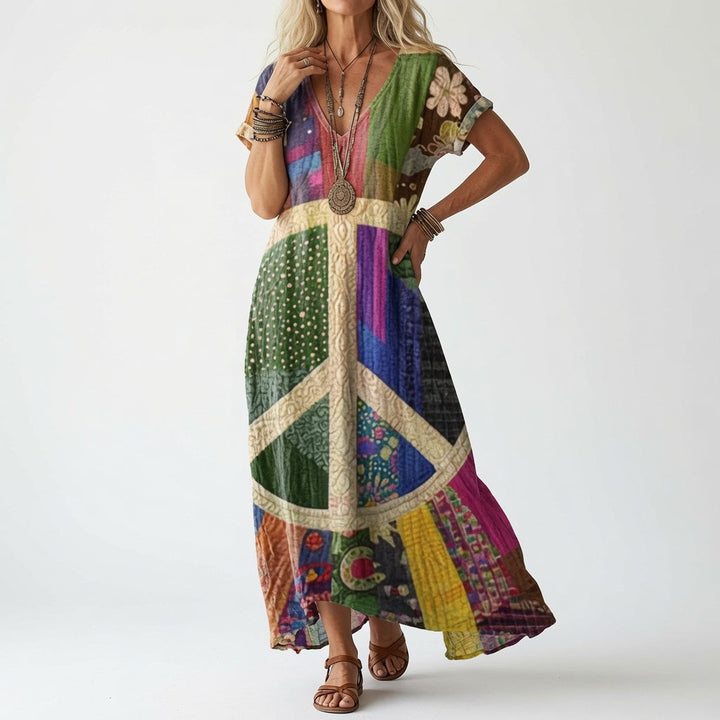 Model draagt de jurk in kleurrijke patchworkstijl met een groot vredesteken in crème op de voorkant, met bruine sandalen en kralenkettingen. 2