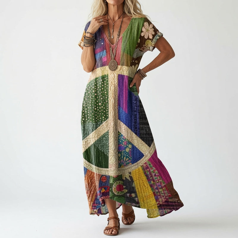 Model draagt de jurk in kleurrijke patchworkstijl met een groot vredesteken in crème op de voorkant, met bruine sandalen en kralenkettingen. 2