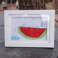 Wit frame op een houten tafel buiten, met een kindertekening van een grote watermeloen met mensjes, ladder en flatgebouw erin. 21