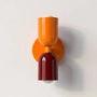 Wandlamp met een oranje bovenkap en een donkerrode onderkap op een oranje ronde wandplaat, tegen een lichte muur. 25