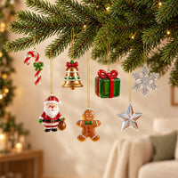 Zeven glazen kersthangers aan gouden draadjes onder een dennentak: zuurstok, klok, kerstman, peperkoekman, cadeautje, ster en sneeuwvlok, met kerstverlichting o 1