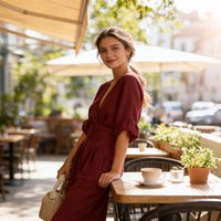 Glimlachende vrouw in donkerrode jumpsuit leunt tegen een tafeltje op een zonnig terras met een kopje koffie. 9