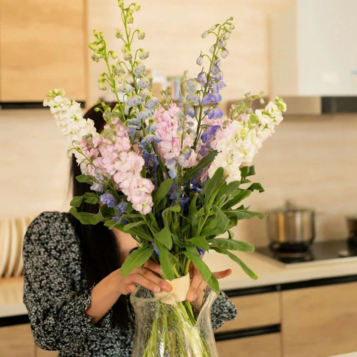 Vrouw in een keuken houdt een hoog boeket roze, witte en blauwe bloemen rechtop in een glazen vaas met de inzet. 2