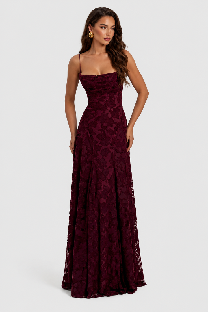 Model in bordeauxrode jurk met reliëfbloemen, gedrapeerde halslijn en dunne bandjes, vooraanzicht met gouden oorbel. 9