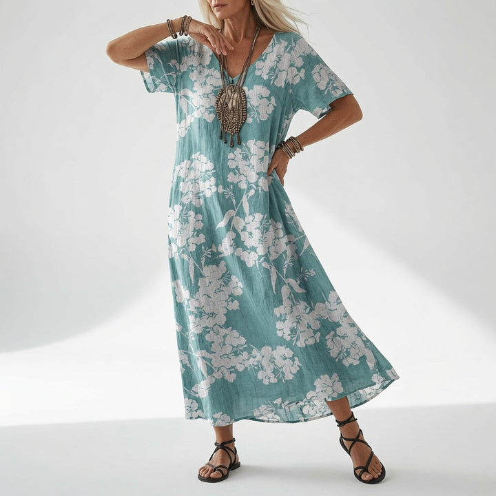 Model draagt de jurk in turquoise met een witte bloesemprint over de hele jurk, met statement-ketting en zwarte sandalen. 4