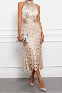 Model draagt de halsjurk in champagnebeige met zilverkleurig metallic clutch, hoge hakken en een rok vol ronde schijfjes. 1