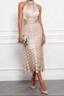 Model draagt de halsjurk in champagnebeige met zilverkleurig metallic clutch, hoge hakken en een rok vol ronde schijfjes. 1