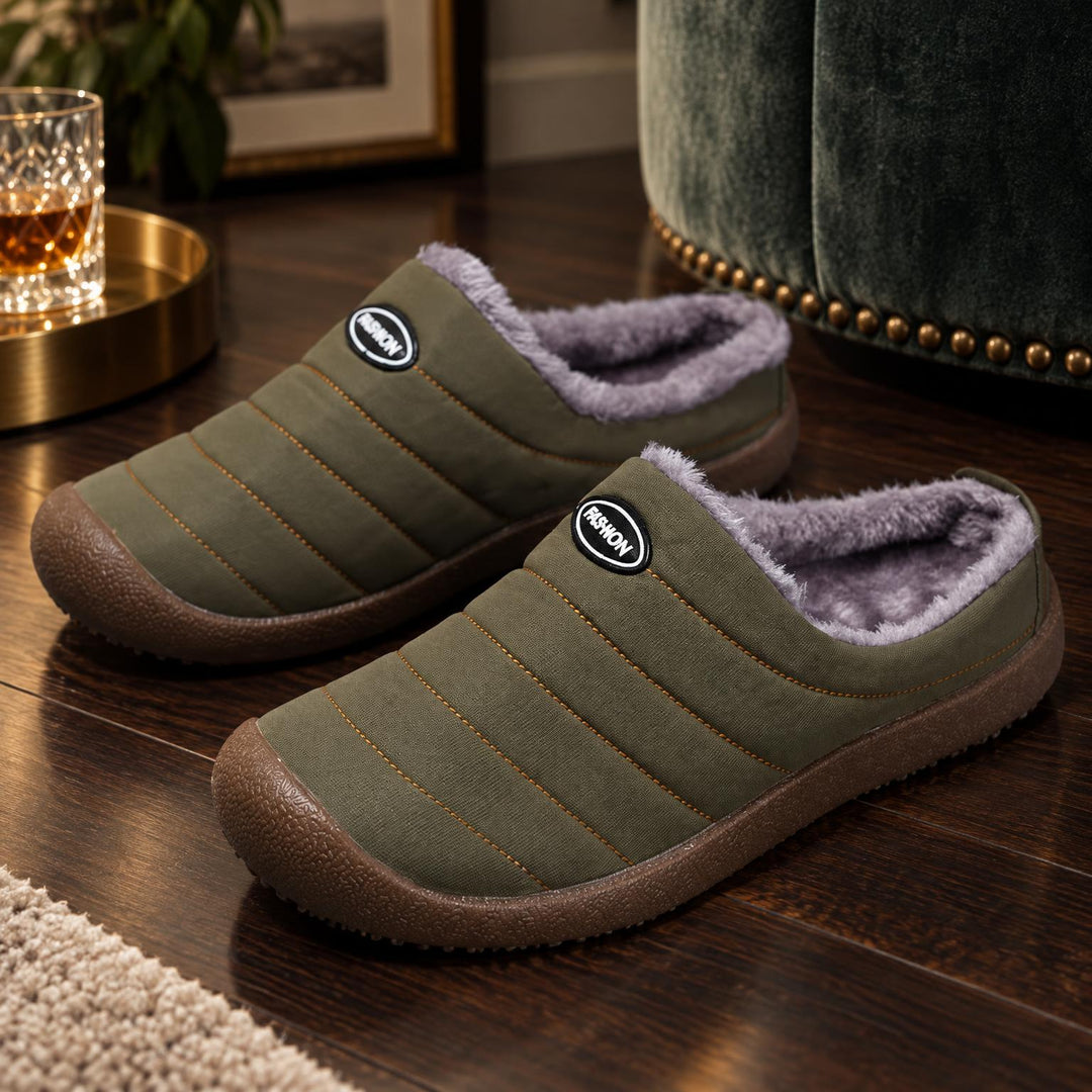 Olijfgroene gewatteerde pantoffels met pluche voering op een houten vloer naast een groene fauteuil 1