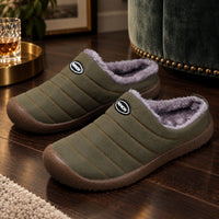Olijfgroene gewatteerde pantoffels met pluche voering op een houten vloer naast een groene fauteuil 1