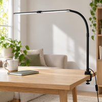 Zwarte klemlamp met gebogen arm en lichtbalk op een houten tafel bij een raam en bank 1