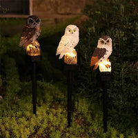 Drie lichtgevende uilen in bruin, wit en bruinwit met oranje vleugels, op grondpinnen tussen lage planten in de avondtuin. 5