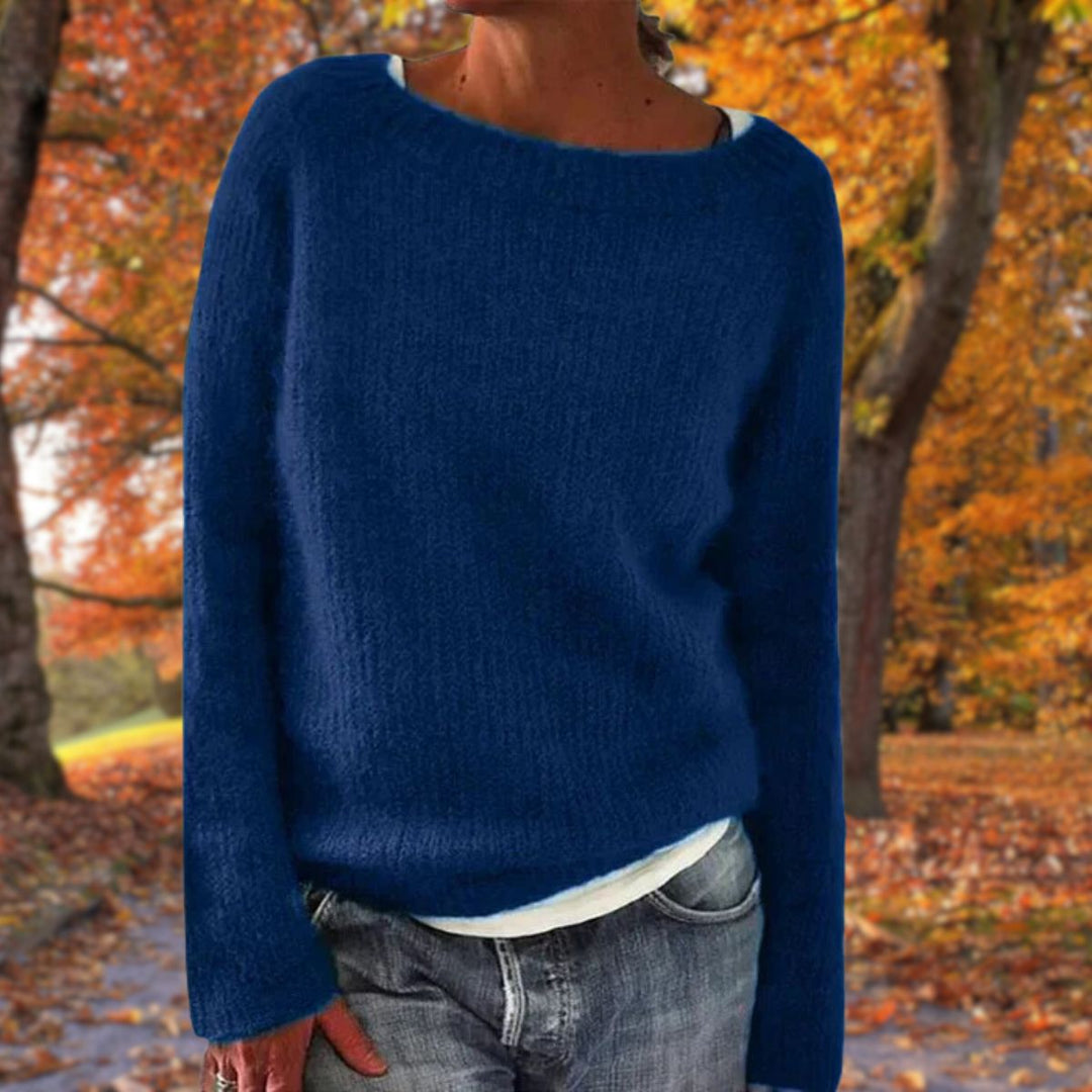 Vrouw draagt de gebreide trui in donkerblauw over een wit shirt en spijkerbroek, met herfstbladeren op de achtergrond. 3