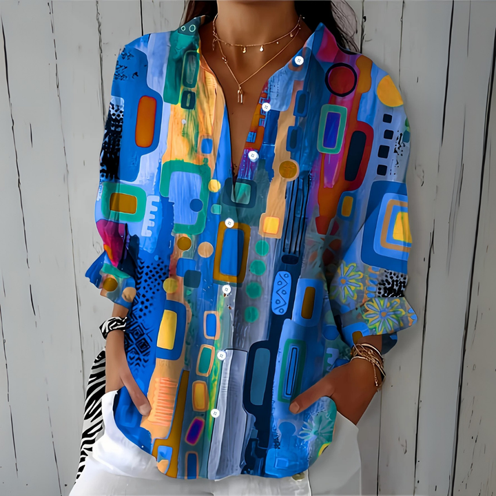 Model draagt de blouse in blauwtinten met kleurrijk abstract patroon, handen in de zakken van een witte broek, voorkant. 1