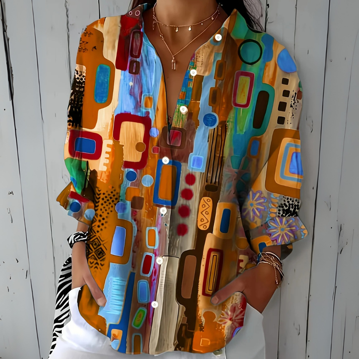 Model draagt de blouse in oranje en lichtblauwe tinten met abstract patroon, losse pasvorm en handen in de zakken. 3