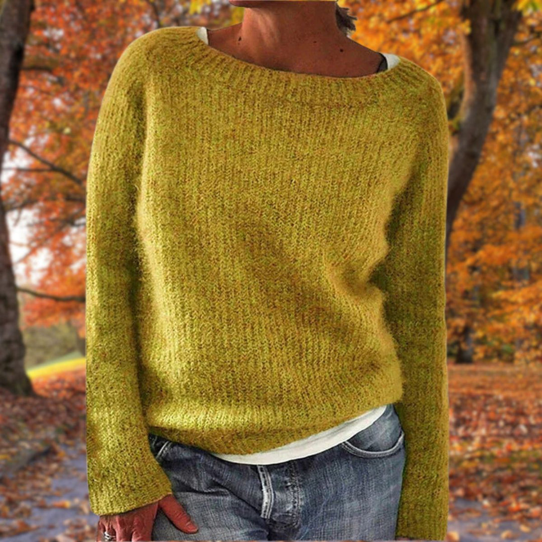 Vrouw draagt de gebreide trui in okergeel over een wit shirt en spijkerbroek, met herfstbomen op de achtergrond. 1