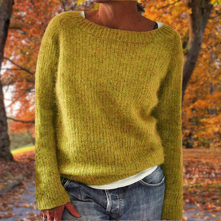 Vrouw draagt de gebreide trui in okergeel over een wit shirt en spijkerbroek, met herfstbomen op de achtergrond. 1