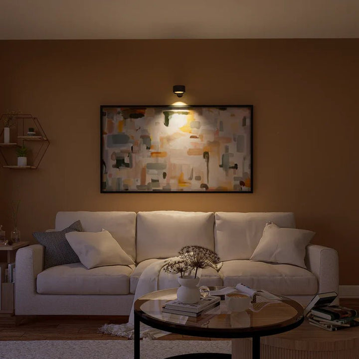 Zwarte wandlamp boven een groot abstract schilderij verlicht de kunst, boven een crèmekleurige bank in een sfeervolle woonkamer. 8