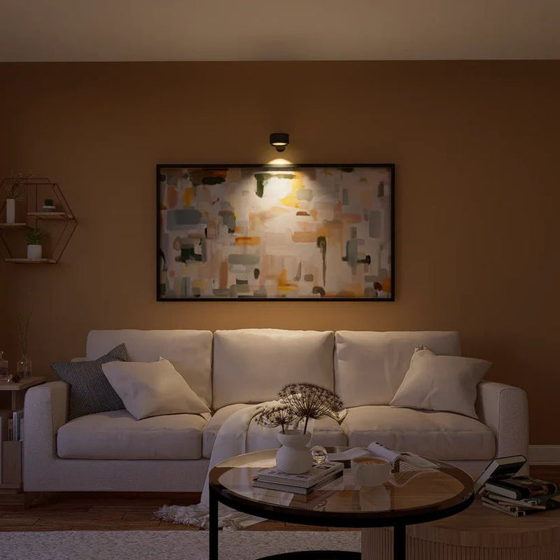 Zwarte wandlamp boven een groot abstract schilderij verlicht de kunst, boven een crèmekleurige bank in een sfeervolle woonkamer. 8