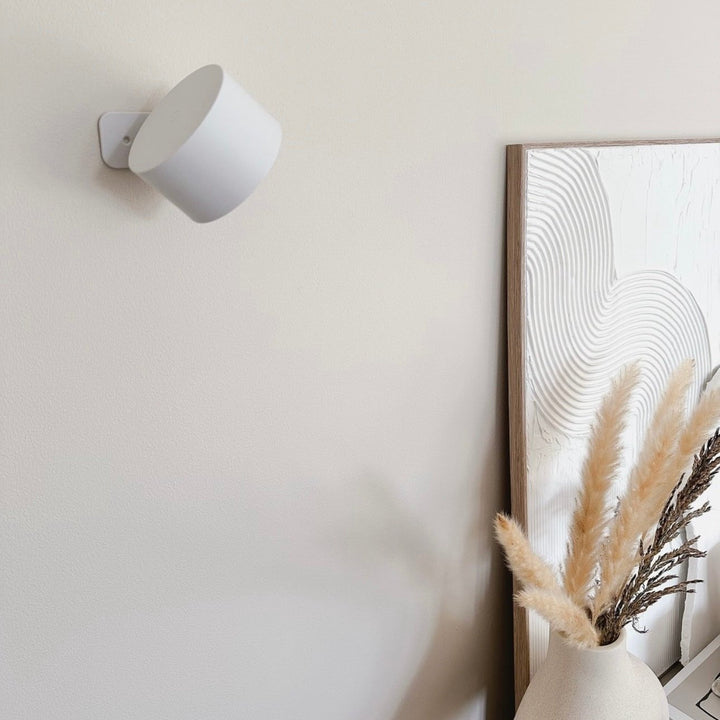 Uitgeschakelde witte wandlamp aan een beige muur, naast een lijst met reliëfschilderij en een vaas met pampasgras. 17