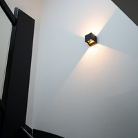 Zwarte kubuslamp aan een witte trapmuur, gezien van onderaf, met warm licht omhoog en omlaag. 2