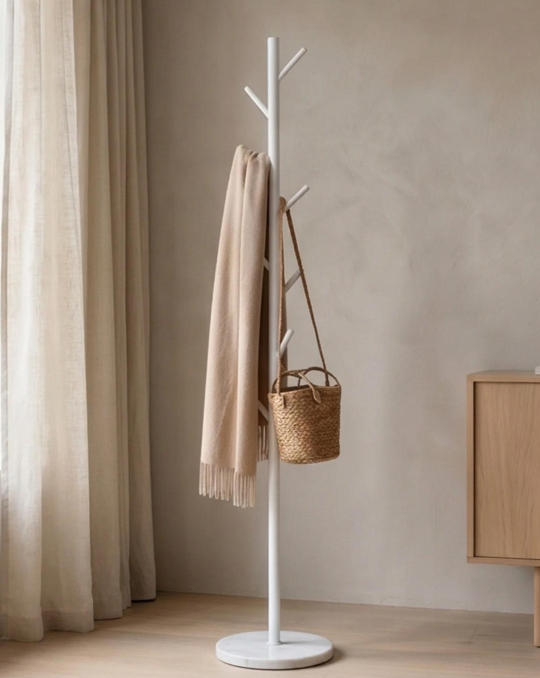 Witte kapstok met een beige sjaal met franjes en een rieten mandtas aan de haken, naast een houten dressoir. 3