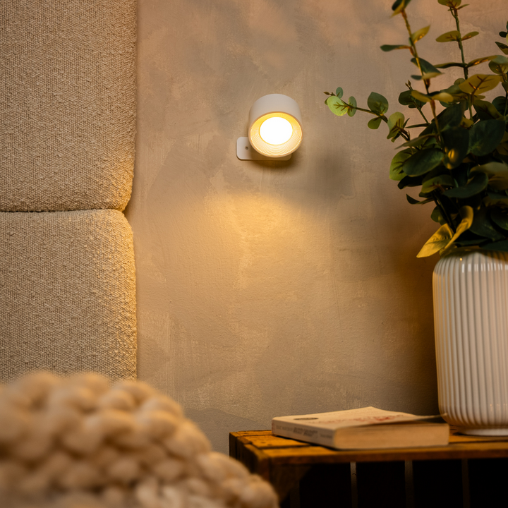 Witte wandlamp brandt met warm licht tegen een gepleisterde muur, naast een vaas met eucalyptus en een boek op een houten kistje. 12