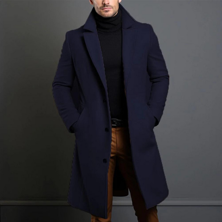 Lange overjas in donkerblauw gedragen door een model met zwarte coltrui en bruine broek, handen in de zakken. 4