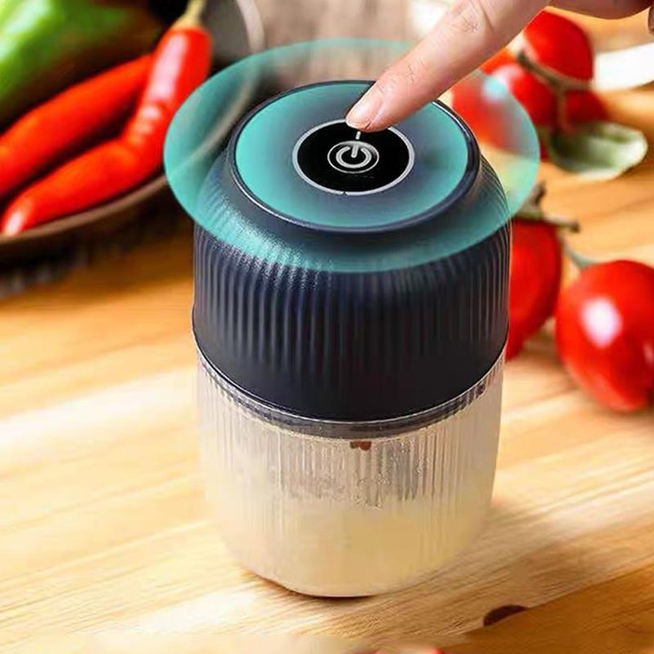 Vinger drukt op de aan-knop bovenop de donkerblauwe hakmolen met geribbelde kom, tussen paprika's en tomaten op een houten plank. 2