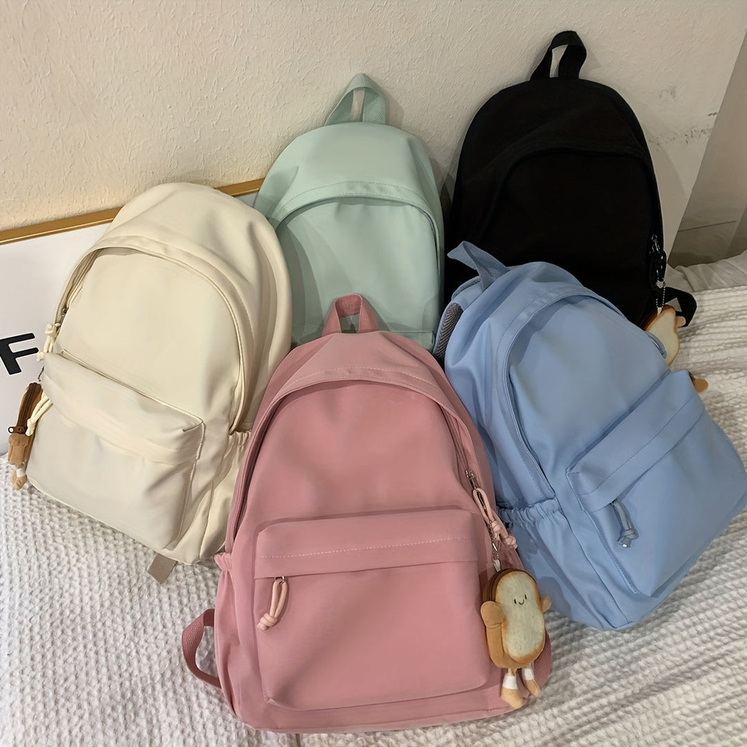 Vijf rugzakken in crème, mint, zwart, roze en blauw samen op een bed, met een broodje-knuffelhanger aan twee tassen. 8