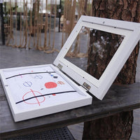 Wit frame geopend op een houten tafel buiten, met een tekening vastgezet onder zwarte elastische banden. 20