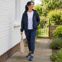 Lachende vrouw in jack en legging loopt met stoffen tas over een tuinpad in blauwe klittenbandschoenen 17