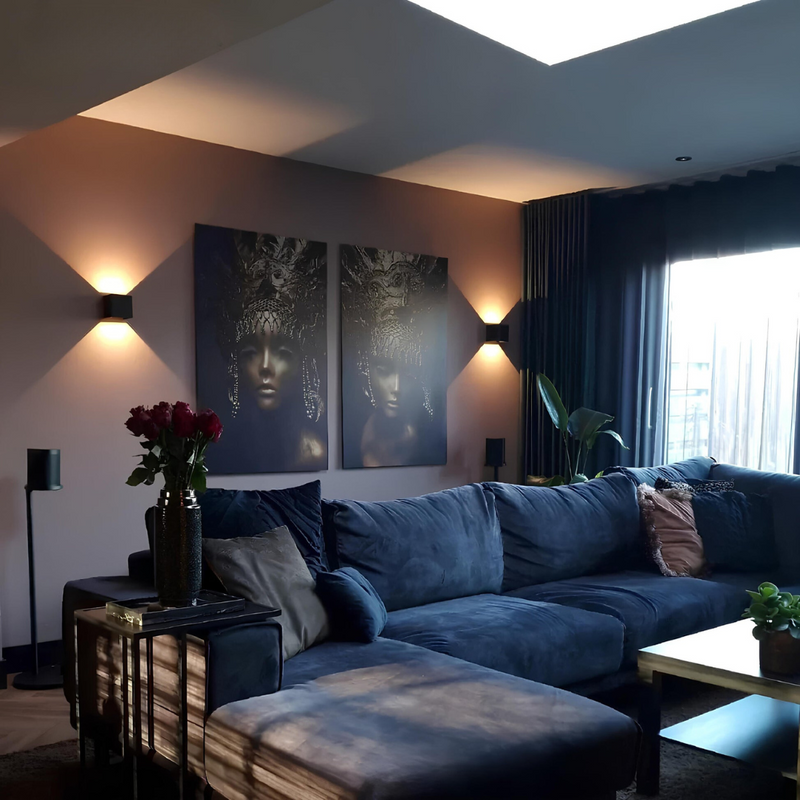 Twee zwarte wandlampen in een woonkamer met blauwe bank, twee portretschilderijen en donkere gordijnen. 9