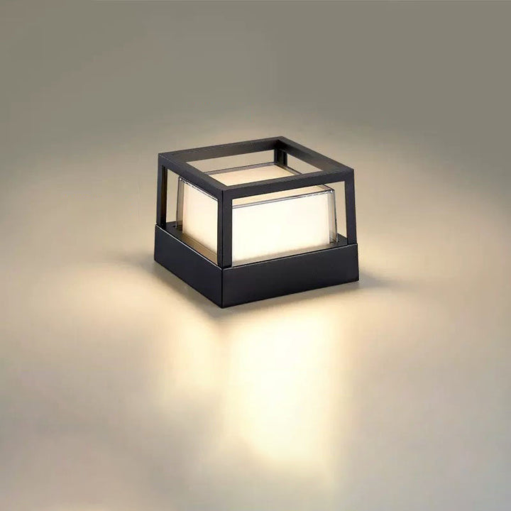 Vierkante zwarte lamp met open kubusvormig frame bovenop en warm licht op een neutrale achtergrond. 11