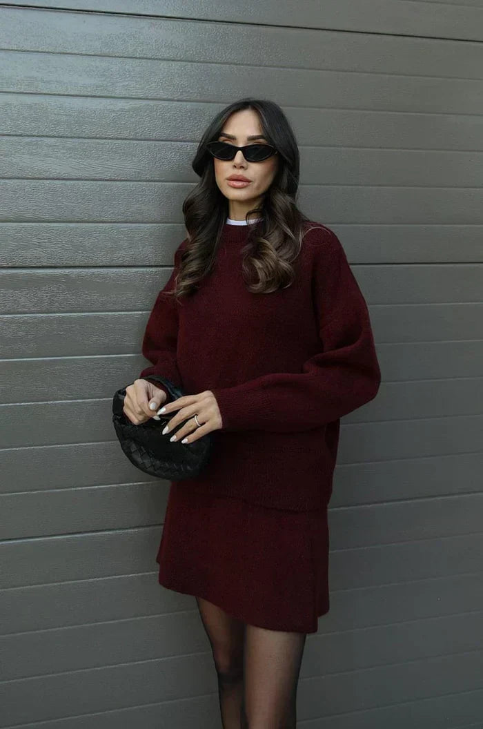Model in bordeauxrode trui en rok, zijaanzicht met gevlochten zwarte tas in beide handen. 3